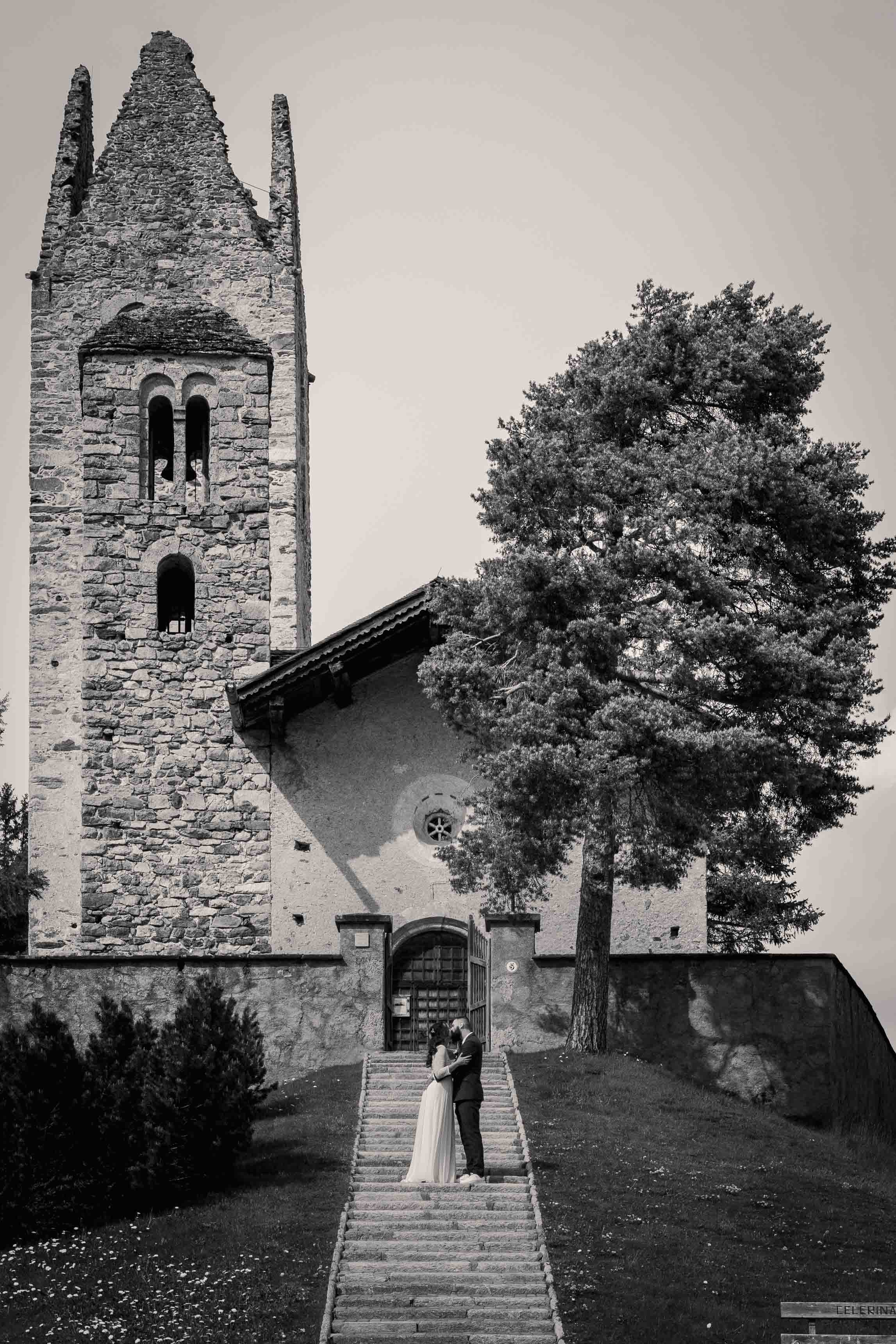 Schwarz-Weiß-Bild eines Paares auf Steintreppe vor einer alten Kirche mit Glockenturm; Baum im Hintergrund.