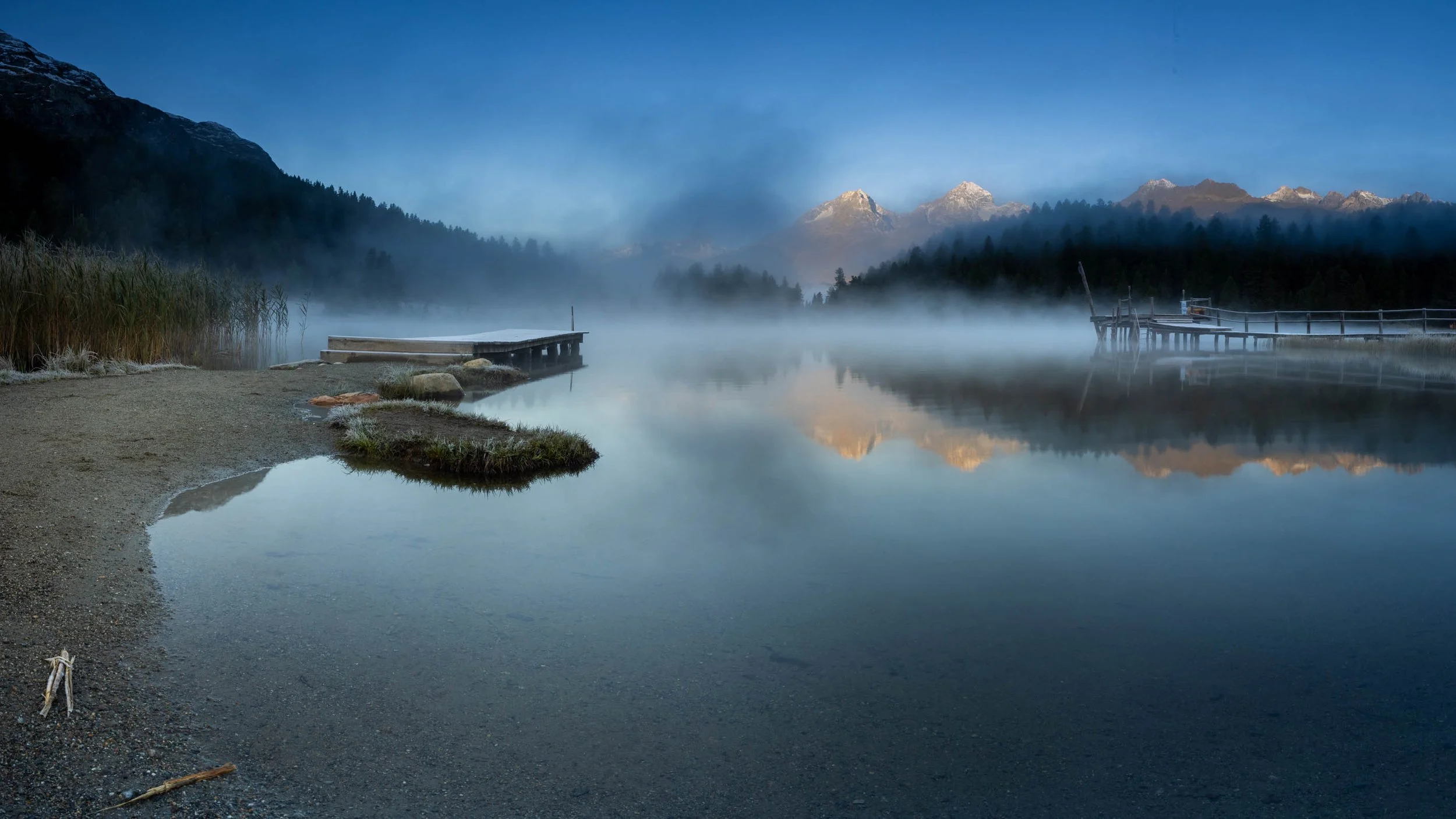 Steg am Stazersee im Morgennebel mit Spiegelung der Engadiner Berglandschaft.