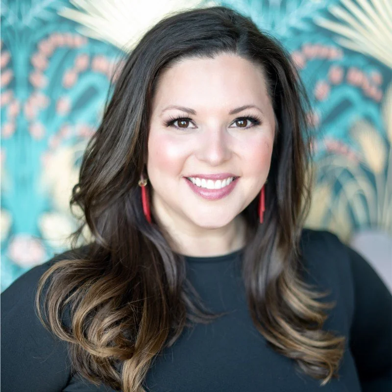 A woman with long, wavy brown hair smiling at the camera, wearing a black top and red earrings, with a colorful, tropical-themed background.