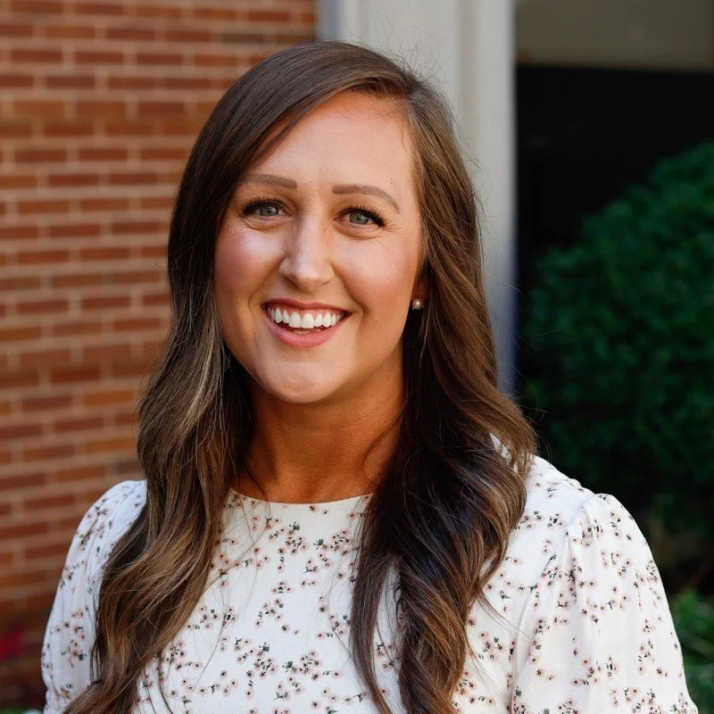 A woman with long brown hair and blue eyes smiling outdoors, wearing a white floral blouse, standing in front of a brick wall and green bushes.