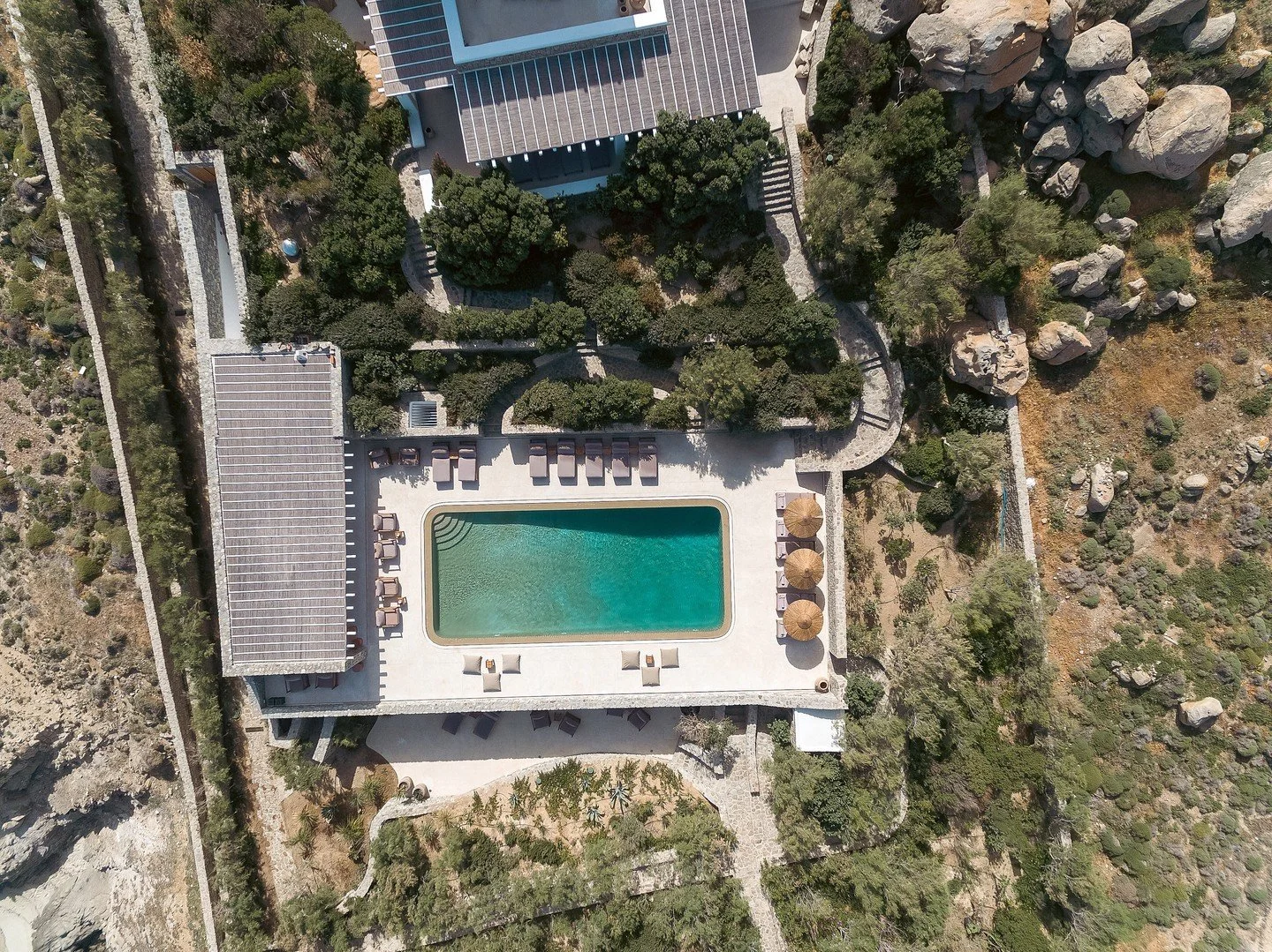 Vue aérienne d'une piscine rectangulaire avec des parasols en paille et des chaises longues autour, entourée de végétation et de rochers.