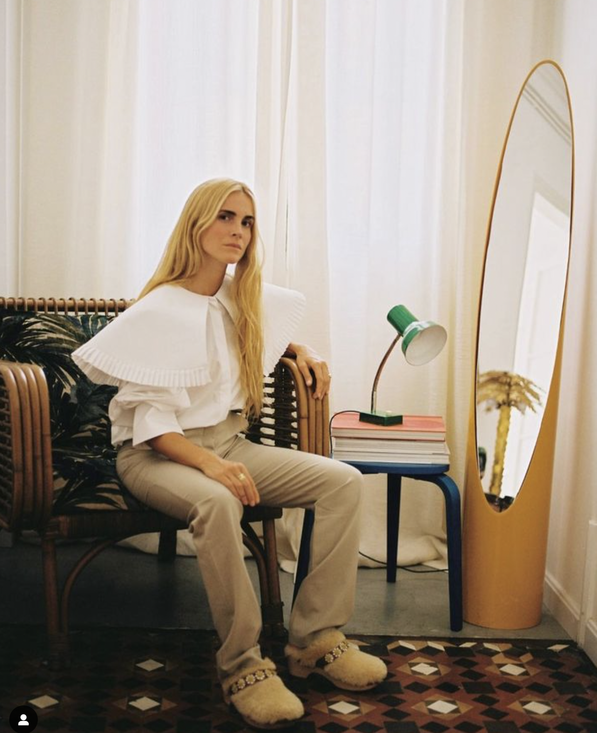 Une femme blonde assise sur un canapé en rotin, portant une chemise blanche et un pantalon beige, dans une pièce lumineuse avec des rideaux clairs, un miroir ovale, et une petite table avec des livres et une lampe