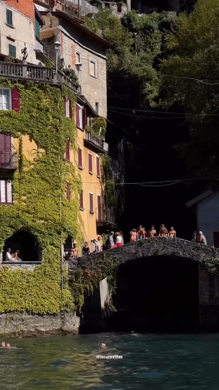 Des personnes se baignent dans une rivière au pied d'une maison ancienne à l'extérieur, avec des murs recouverts de verdure et un petit pont en pierre.
