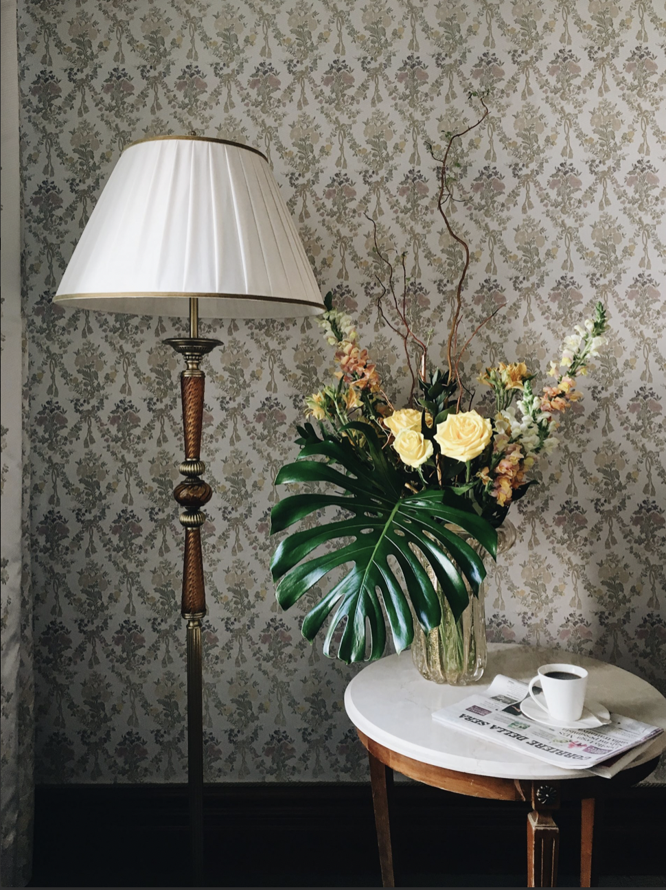 Une lampe de lecture à côté d’un vase avec des fleurs tropicales, une tasse de café et un journal posés sur une petite table en bois.