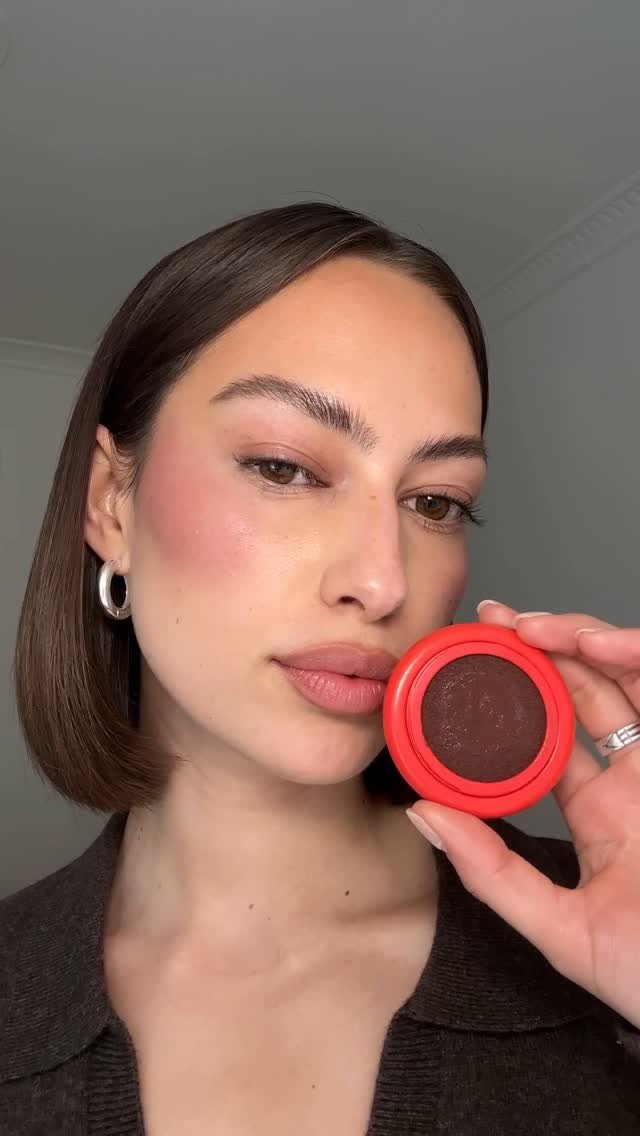 Une femme souriante avec une boucle d'oreille ronde en argent, tenant un petit pot de maquillage rouge, probablement du rouge à lèvres ou de la blush, devant un mur gris clair.