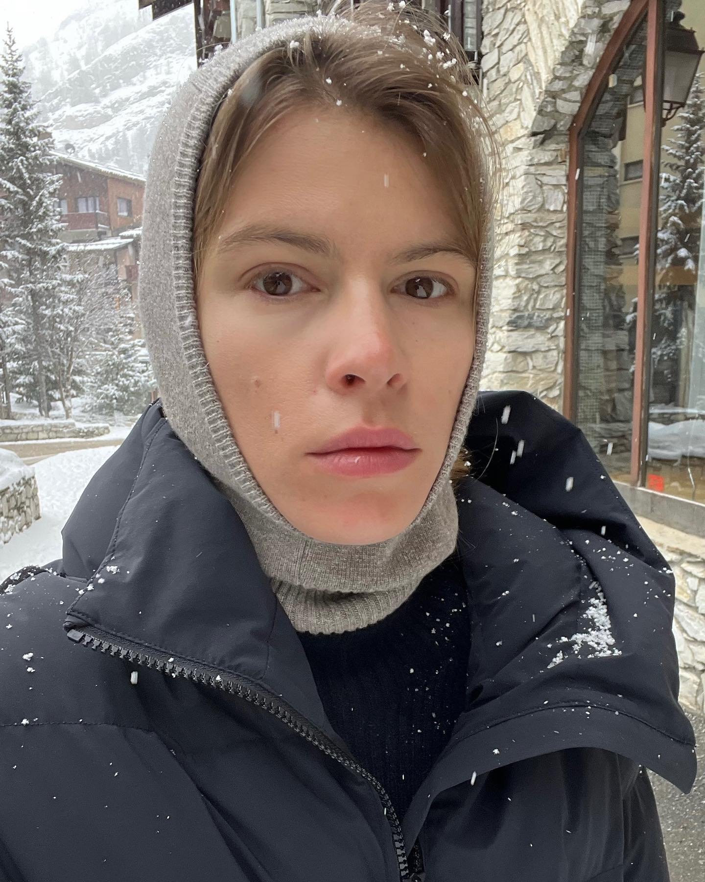 Jeune femme en plein air par temps neigeux, portant un bonnet gris, un pull en laine gris et une veste noire, devant un bâtiment en pierre avec des fenêtres en verre.