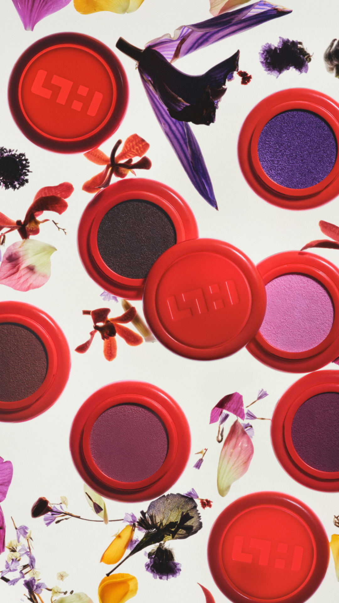 Divers pots de maquillage avec ombres à paupières colorées, entourés de fleurs séchées et de pétales, sur un fond blanc.