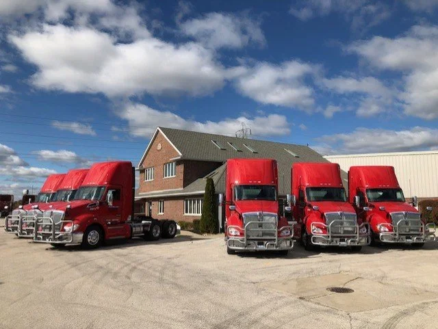 Four red semi-trucks parked in front of a brick building under a partly cloudy sky.