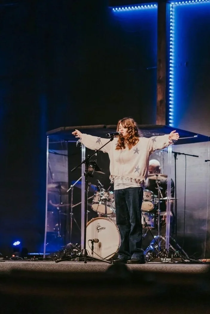 A woman singing on stage with her arms outstretched, behind a drum set, with stage lights and equipment around her.