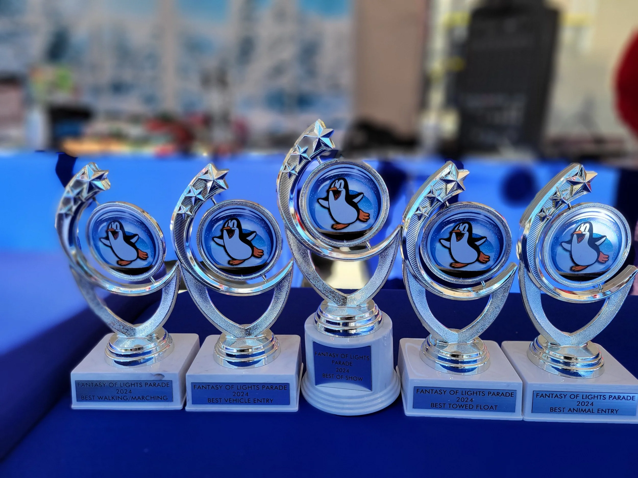 Six trophies with a penguin logo, awarded at the Fantasy of Lights Parade 2024 for categories including best walking/marching, best vehicle entry, best of show, best towed float, and best animal entry, displayed on a table.