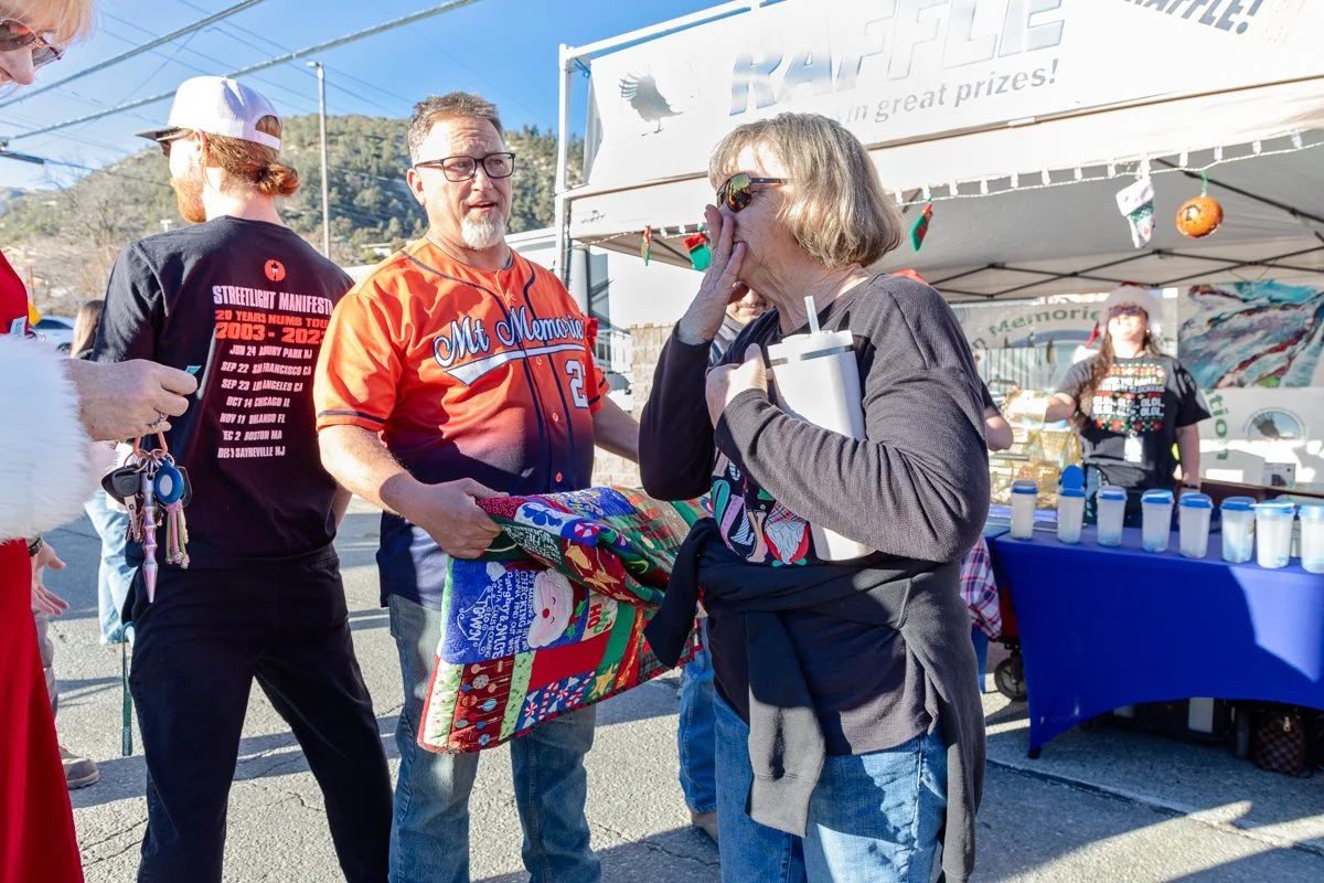 People at an outdoor holiday market, with a woman holding a gift and an elderly woman reacting surprised or emotional. There is a booth with candles and holiday decorations in the background.