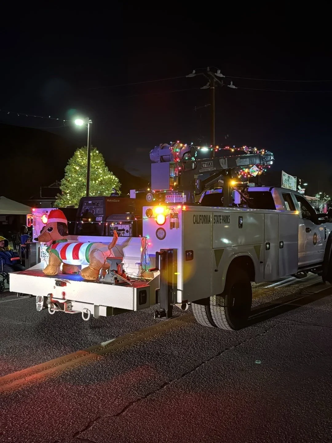 A decorated California State Parks utility truck with colorful holiday lights and a large inflatable dog wearing a Santa hat and festive sweater on the back platform, parked at night in a Christmas parade or festival scene with illuminated trees and