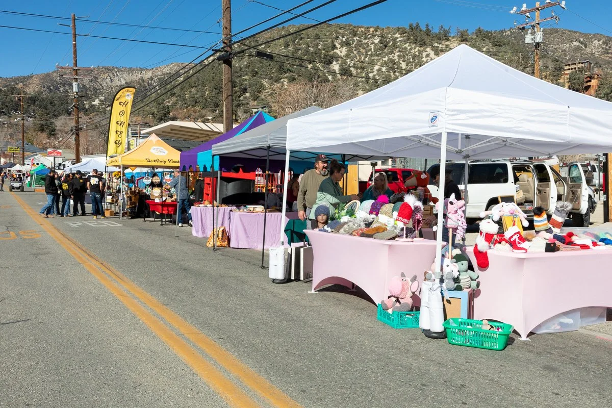 Outdoor street market with vendor tents selling handmade and craft items, people browsing and shopping, in a mountainous area under clear blue sky.