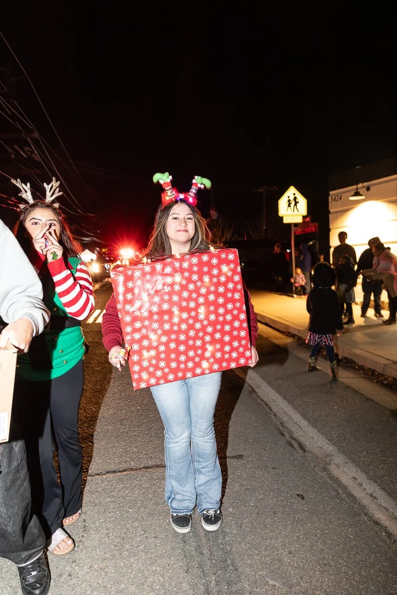 Person dressed in Christmas attire holding a large red gift box with white snowflake patterns during a nighttime holiday event.