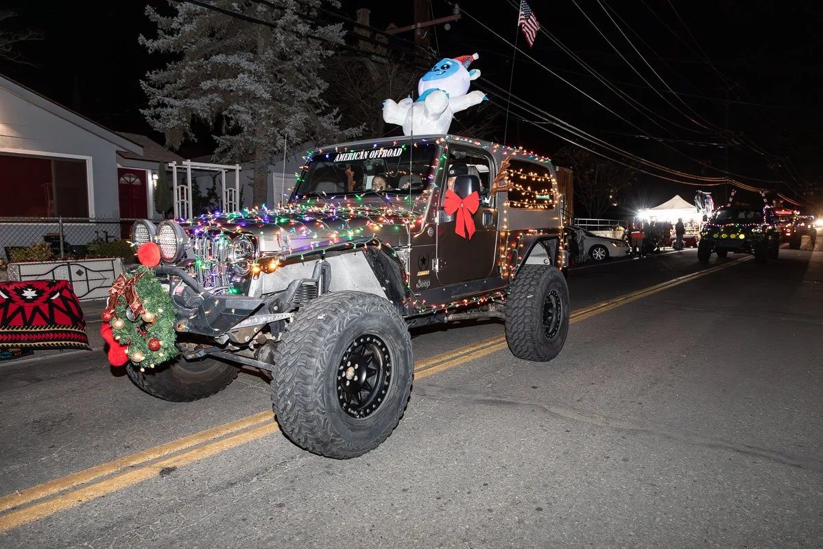 A decorated Jeep with Christmas lights and ornaments, a small Christmas wreath on the bumper, and a large inflatable snowman on the roof, participating in a nighttime holiday parade.