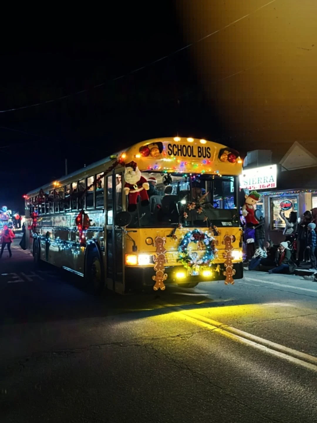Nighttime parade float decorated with Christmas ornaments, lights, and festive decorations, featuring a school bus labeled 'School Bus' at the top, with Santa Claus and reindeer figures, and people watching along the street.
