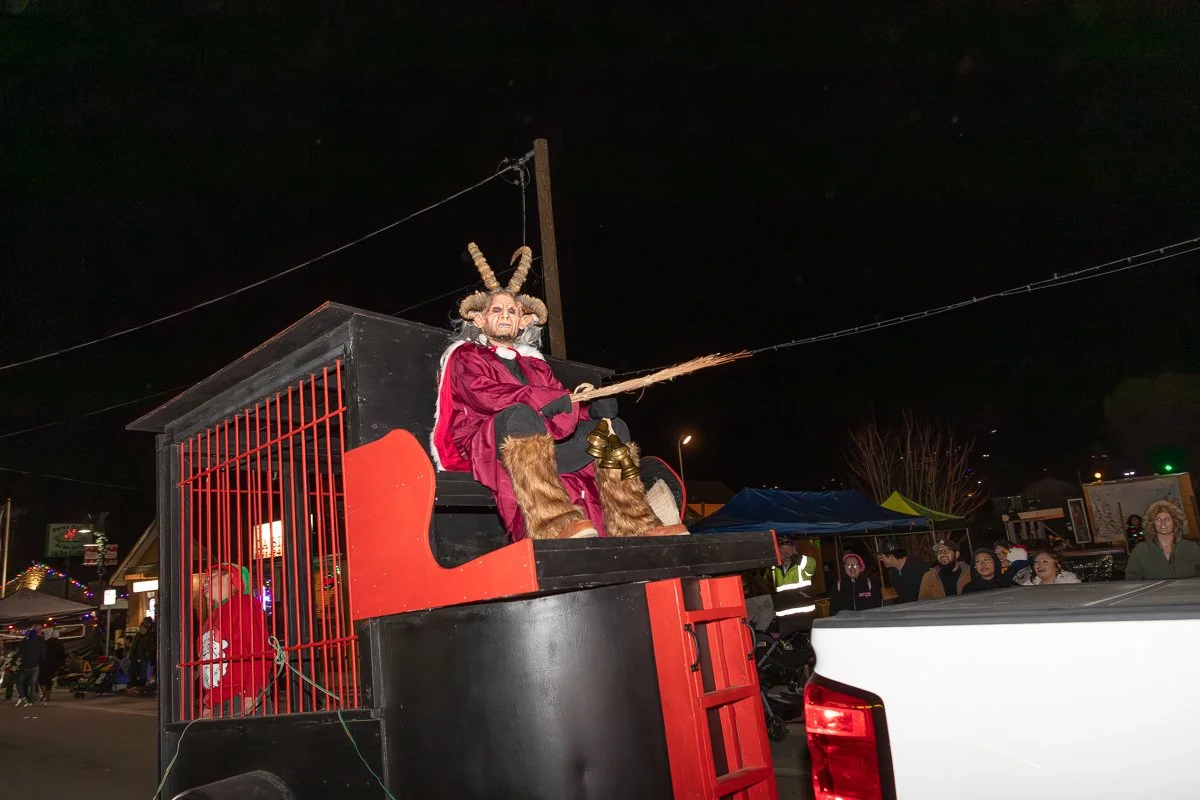 Person dressed as a mythical goat figure with large horns, sitting on a float at night, waving a stick, during a parade or festival.