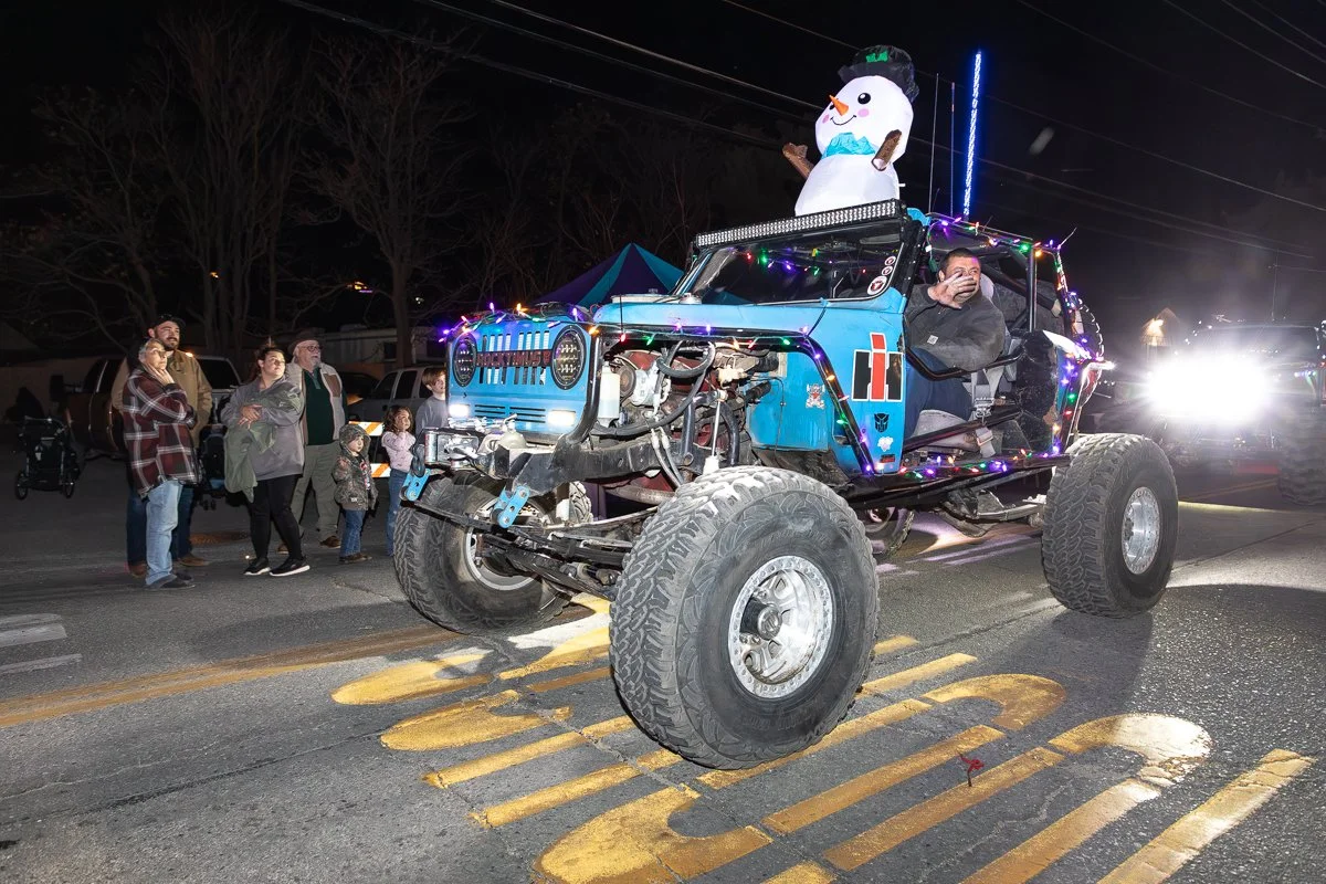 A decorated off-road vehicle with a snowman figure on top, surrounded by people at night in a parade or event, with bright lights and Christmas decorations.
