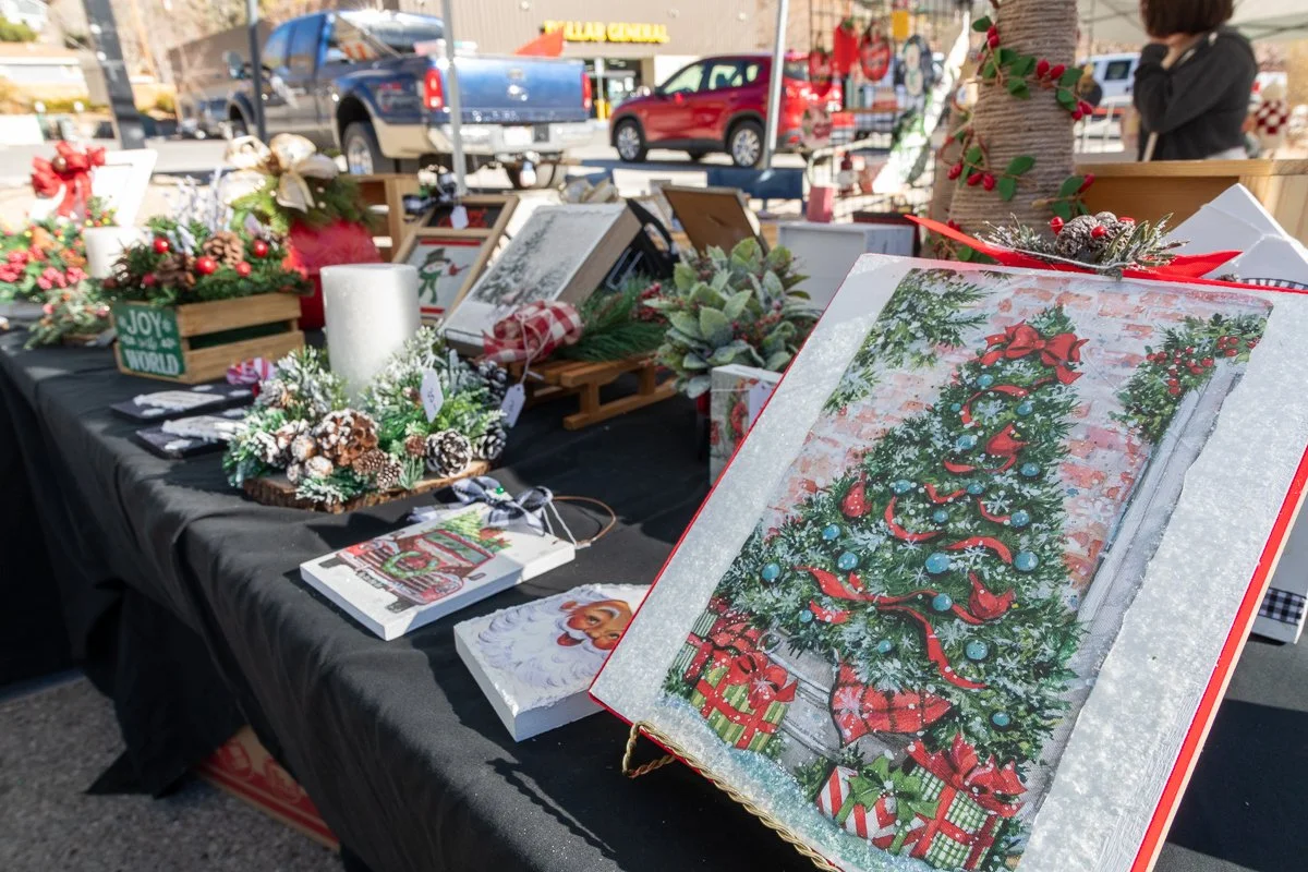 Table at a Christmas market displaying holiday decorations, framed holiday artwork, and Christmas-themed arrangements, with a Christmas tree decorated with ornaments and bows in the foreground.