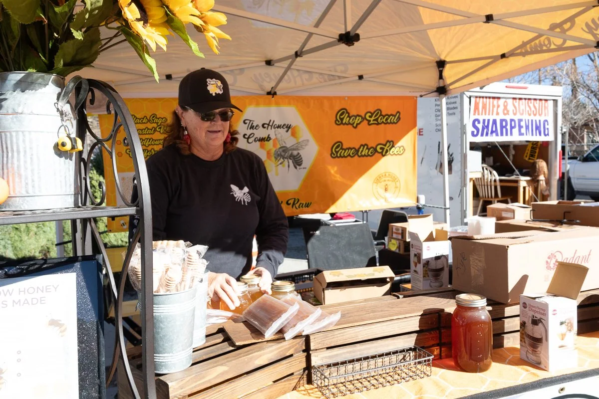 A woman at a honey stand selling honey products at an outdoor market. She is wearing a black hat and sunglasses, and there are jars of honey on the table. A yellow honey banner and other market stalls are in the background.
