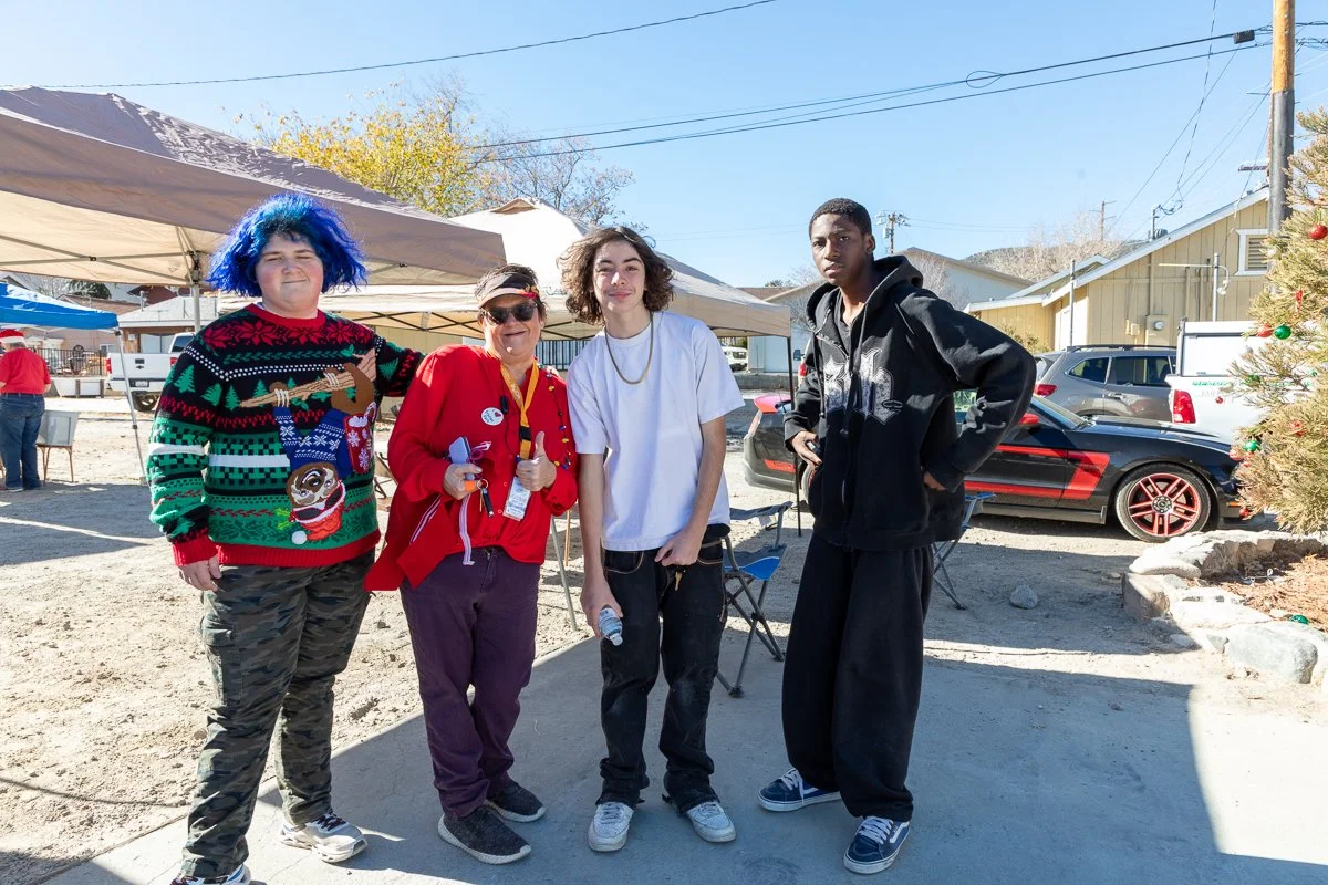 Four people standing outdoors in a sunny area, with tents, cars, and a Christmas tree with ornaments in the background. The person on the far left has blue hair and is wearing a colorful Christmas sweater, the second person is wearing sunglasses and