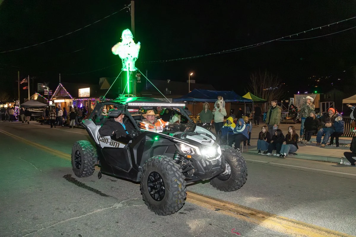 A black and white off-road utility vehicle with a passenger and driver inside, driving down a street at night. The vehicle has a green lightsaber on top, shaped like a snowman, and a crowd of people are sitting and standing along the sidewalk watchin
