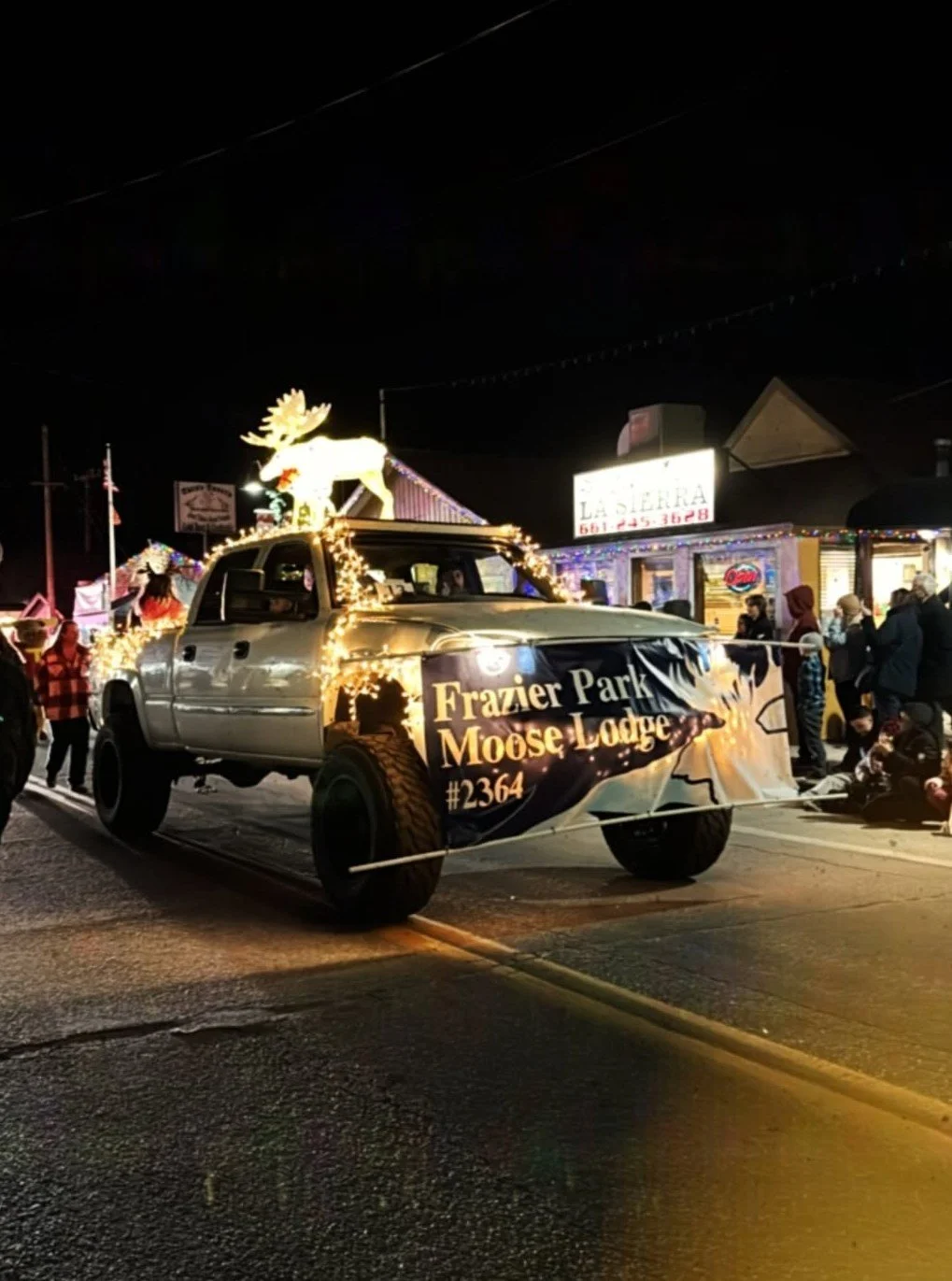 A decorated pickup truck in a nighttime parade, featuring an illuminated moose figure and a banner that reads 'Frazier Park Moose Lodge #2364', with people and a building in the background.