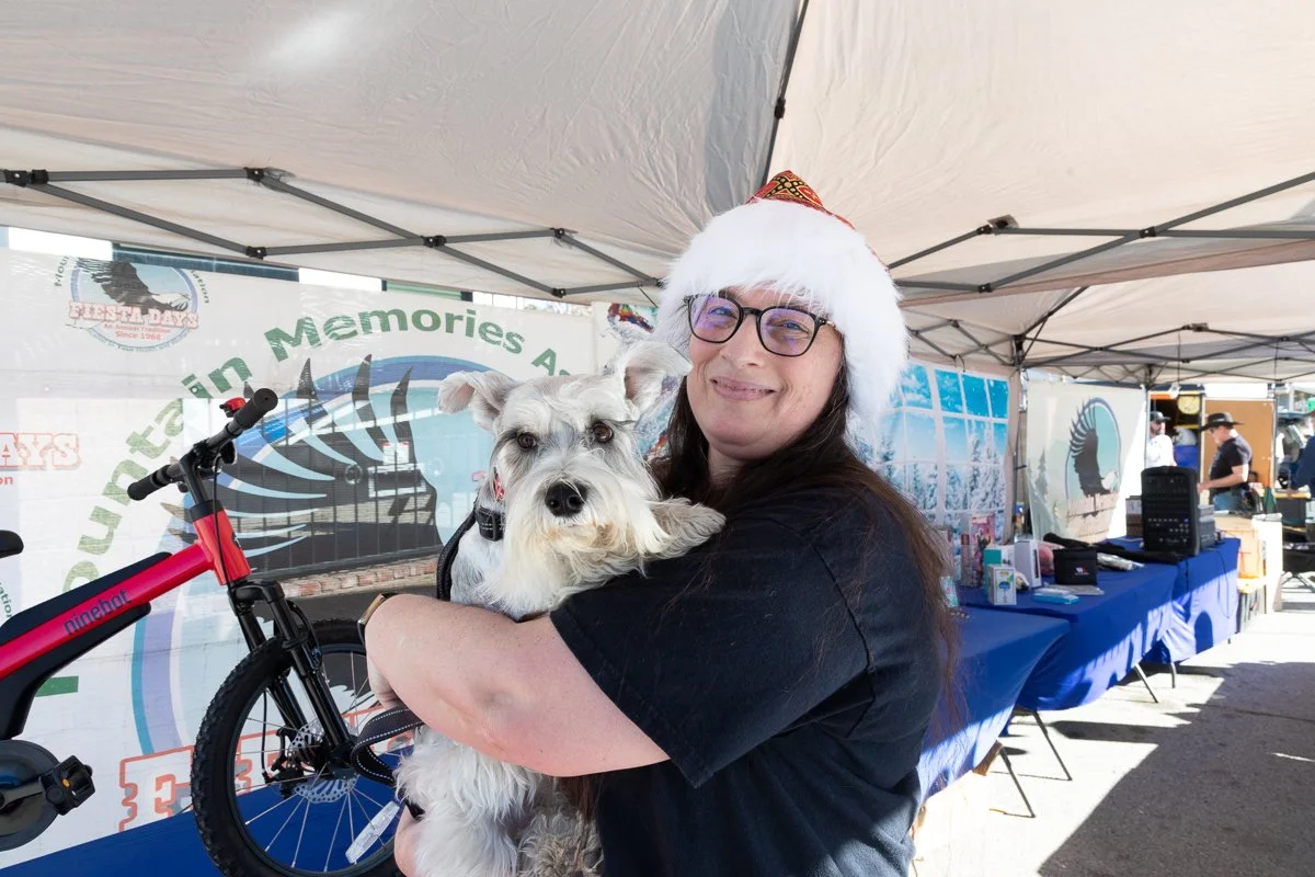 A woman wearing a Santa hat and glasses holding a dog at an outdoor event with a tent and tables in the background.