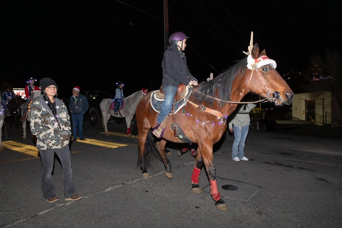 A person riding a brown horse decorated for Christmas with a Santa hat on the horse's head, during a night parade, while a woman stands nearby and other riders on horses are visible in the background with holiday lights.