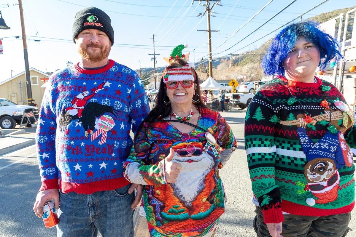 Three people standing outdoors wearing colorful Christmas sweaters, with a street scene and tents in the background.