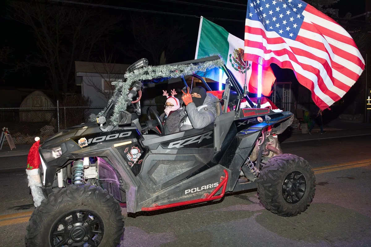 A Polaris RZR off-road vehicle decorated with Christmas decorations, two people wearing masks and holiday accessories inside, and large American and Mexican flags attached to the back, parked on a street at night.