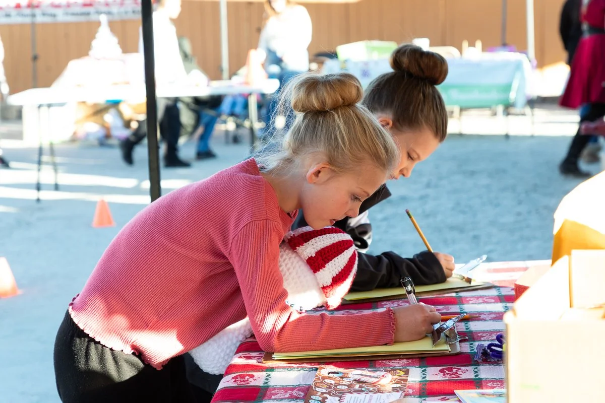 Two young girls are leaning over a table, writing in notebooks at an outdoor event, with one wearing a red sweater and a white and red hat.