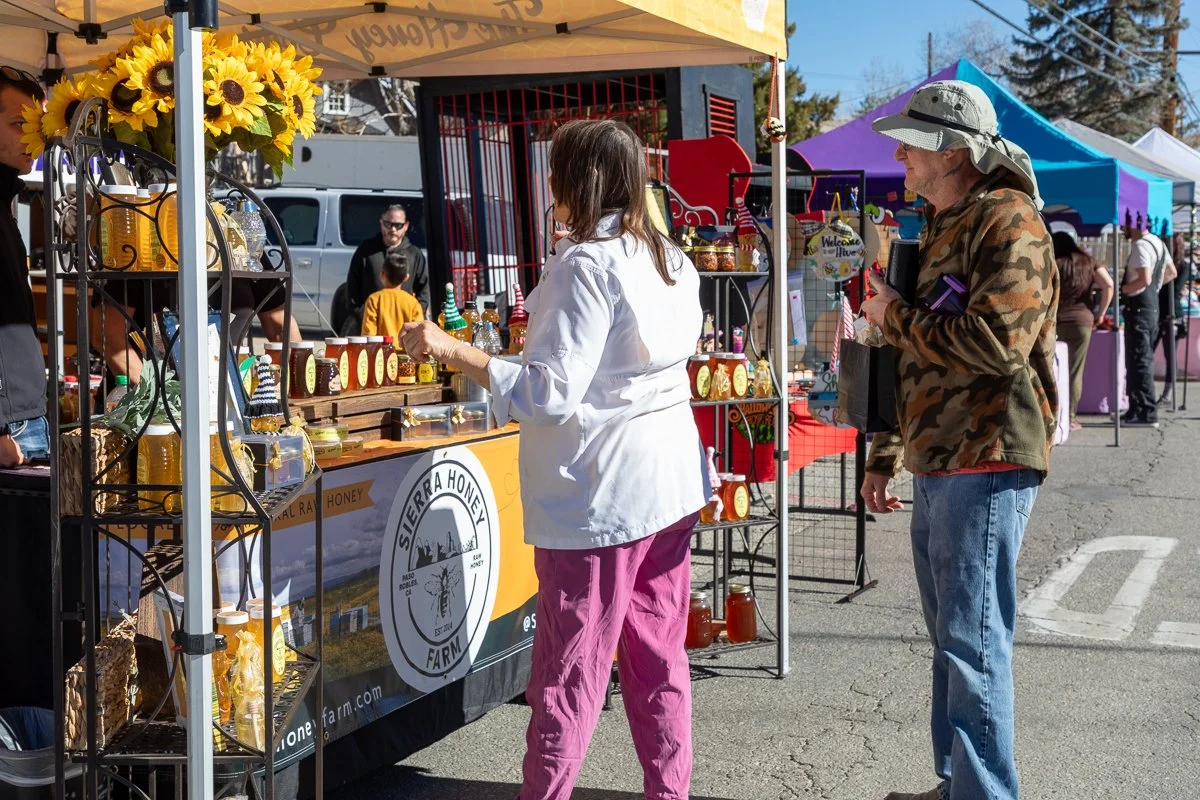 A woman with brown hair wearing a white shirt and pink pants buys honey at a market stall, while a man in a camo jacket and wide-brimmed hat looks on, at an outdoor marketplace with colorful tents and people in the background.