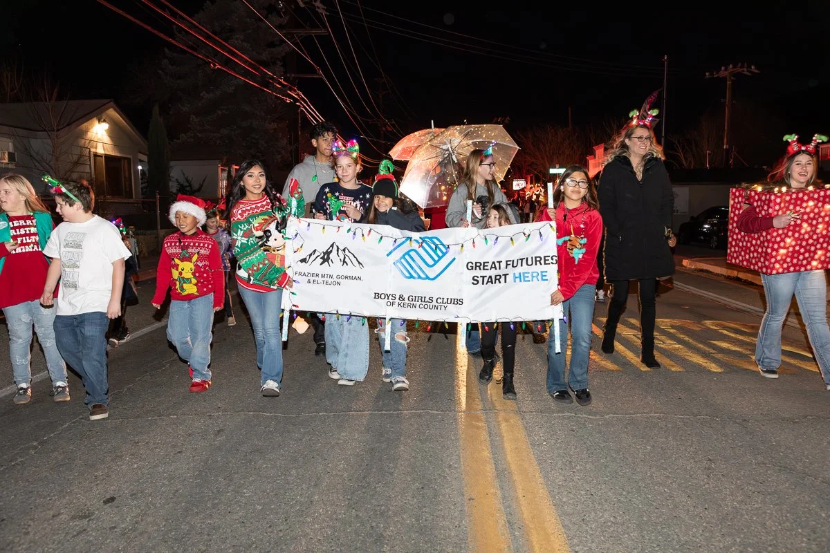 Children and adults participating in a Christmas parade, holding a banner that reads "Great Futures Start Here," and wearing holiday-themed clothing and accessories, with festive lights and houses in the background.