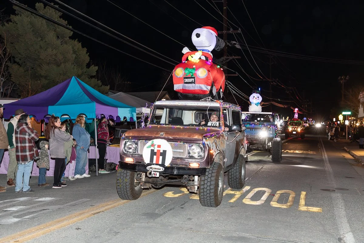 Nighttime parade with decorated vehicles, including a Snoopy float with large Snoopy figure on top, and a crowd of spectators lining the street.