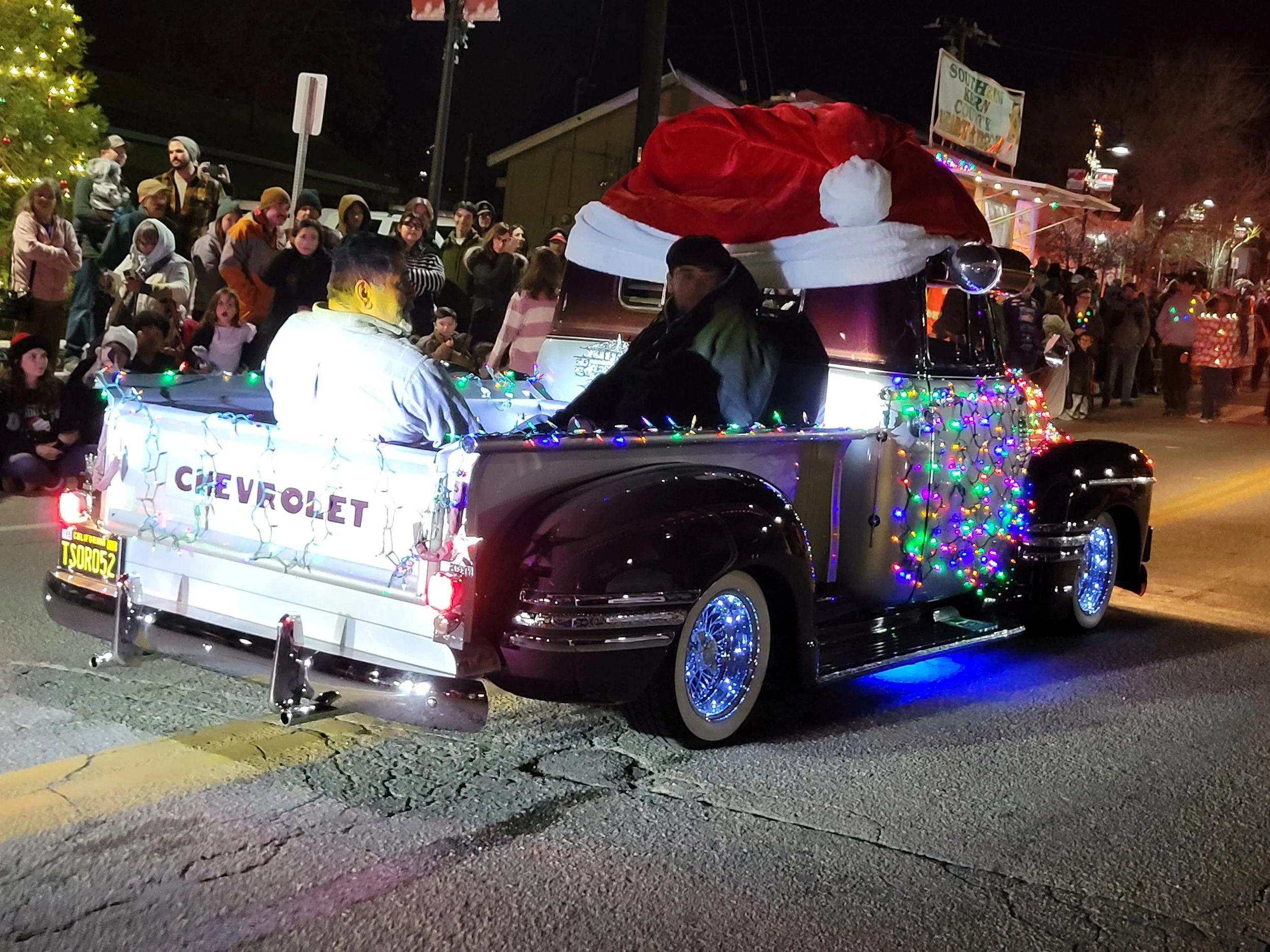 A decorated vintage Chevrolet truck with Christmas lights and a large Santa hat on top. The truck is filled with people and has a sign that reads 'CHEVROLET' on the back. It is part of a nighttime holiday parade with a crowd of spectators gathered al