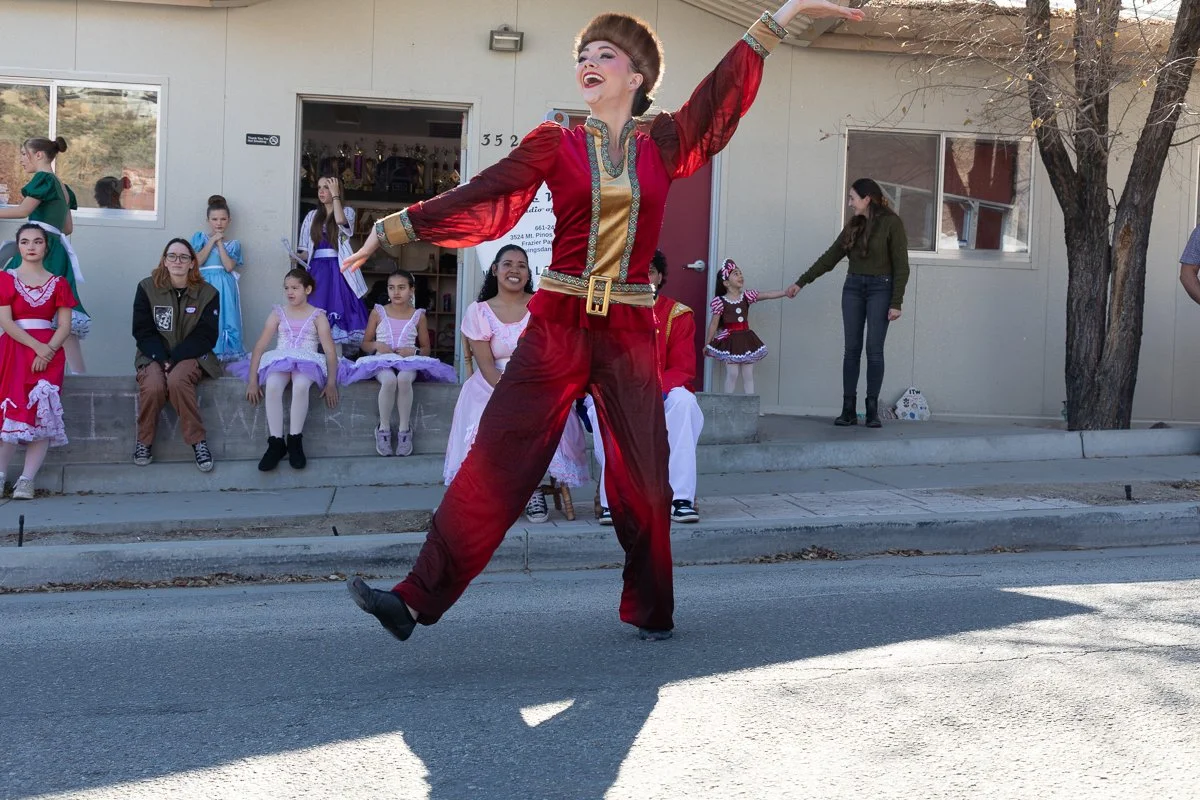 A woman in a colorful traditional costume is performing a dance on the street, with children dressed in various colorful costumes sitting and watching in the background.
