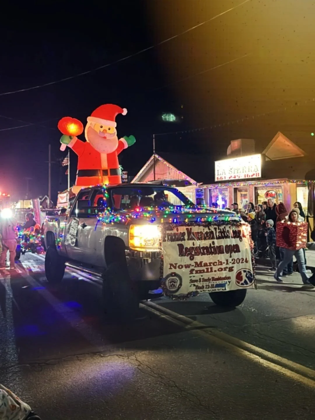 A decorated pickup truck with Christmas lights and a large inflatable Santa Claus on top, participating in a nighttime parade with people walking and carrying gifts.