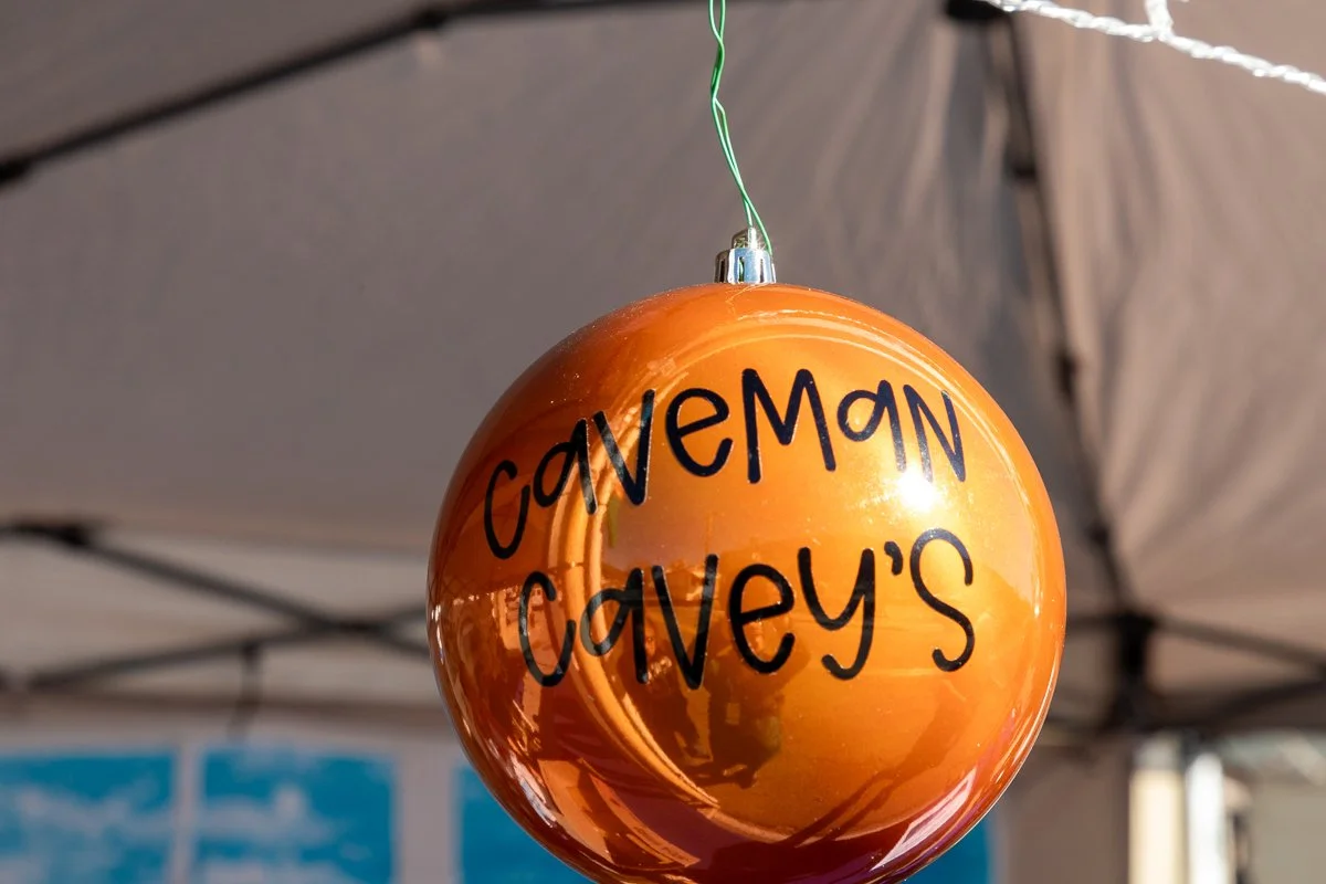 Orange Christmas ornament with black text reading 'Caveman Cavey's' hanging from a green string under a canopy.