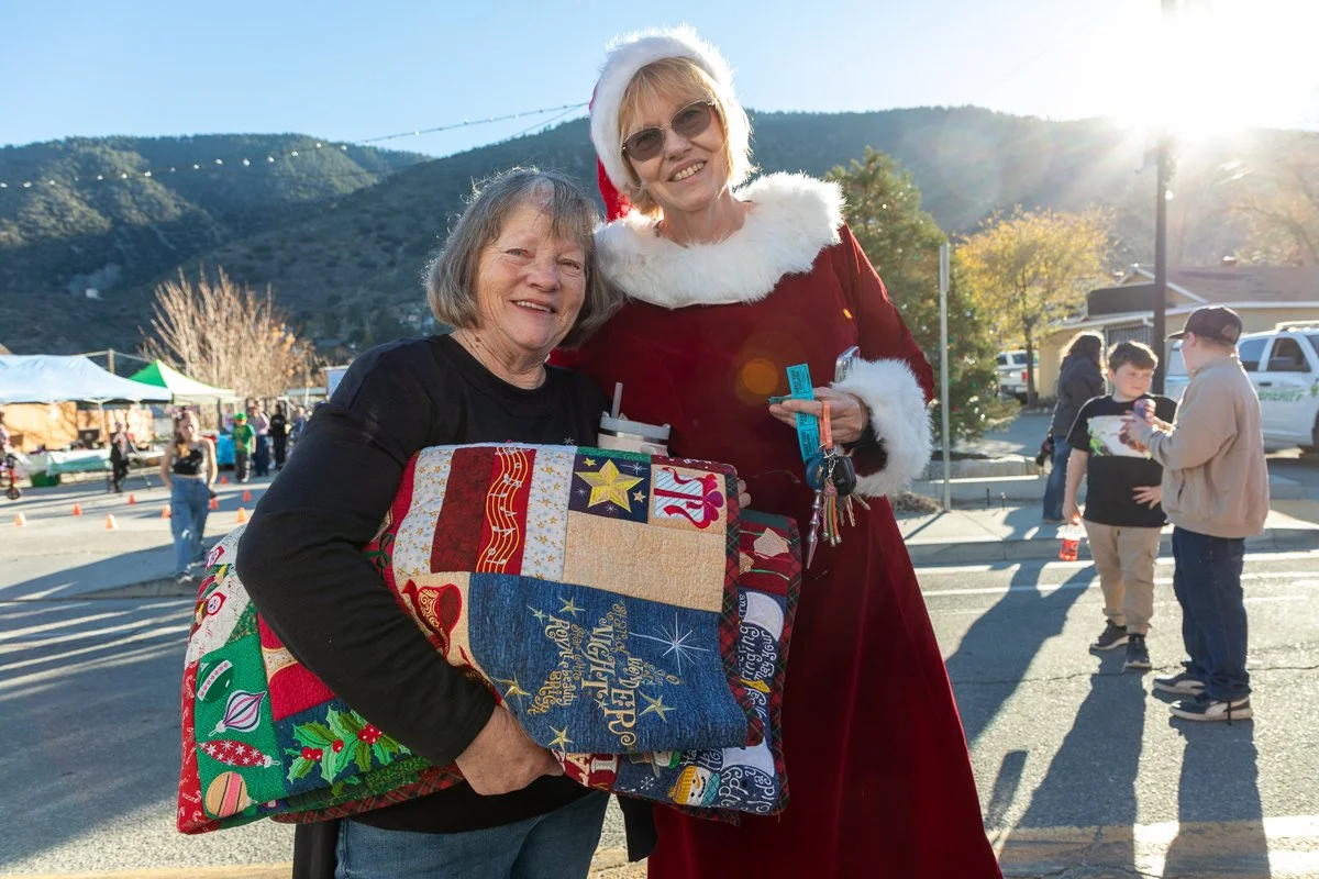 A woman and a sign language interpreter dressed in a Santa costume posing with a woman holding a Christmas-themed gift bag at an outdoor holiday event.