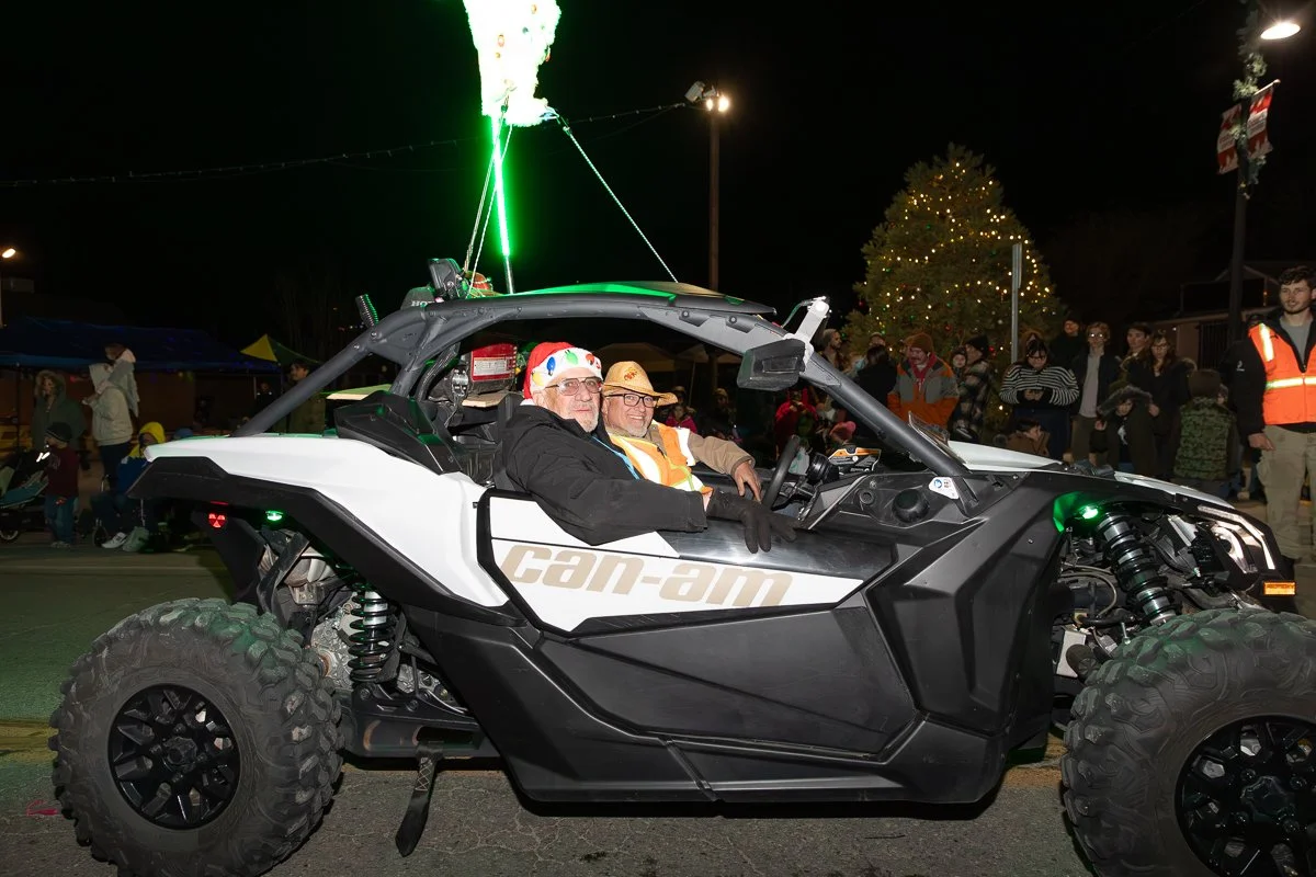 A white and black Can-Am off-road vehicle with two men inside, wearing festive holiday hats, at a nighttime Christmas event with a decorated Christmas tree and people in the background.