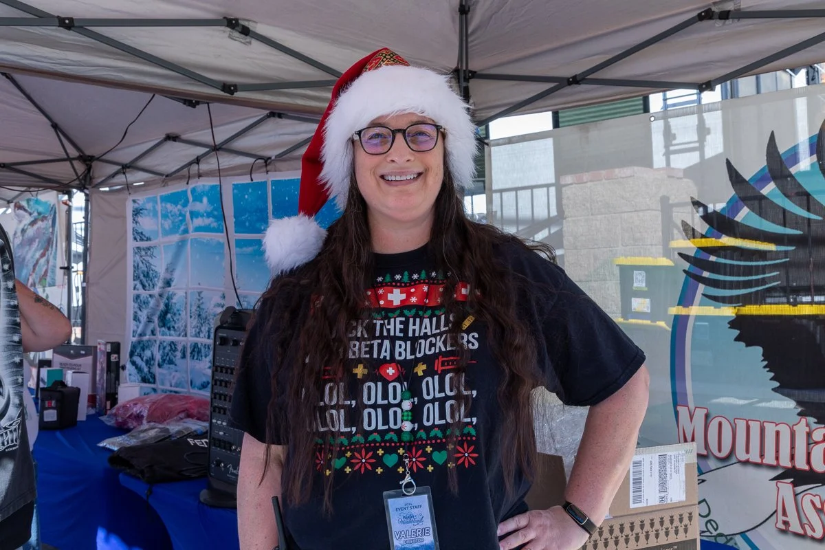 Woman smiling, wearing a Santa hat and Christmas-themed T-shirt, standing under a canopy at an outdoor event.