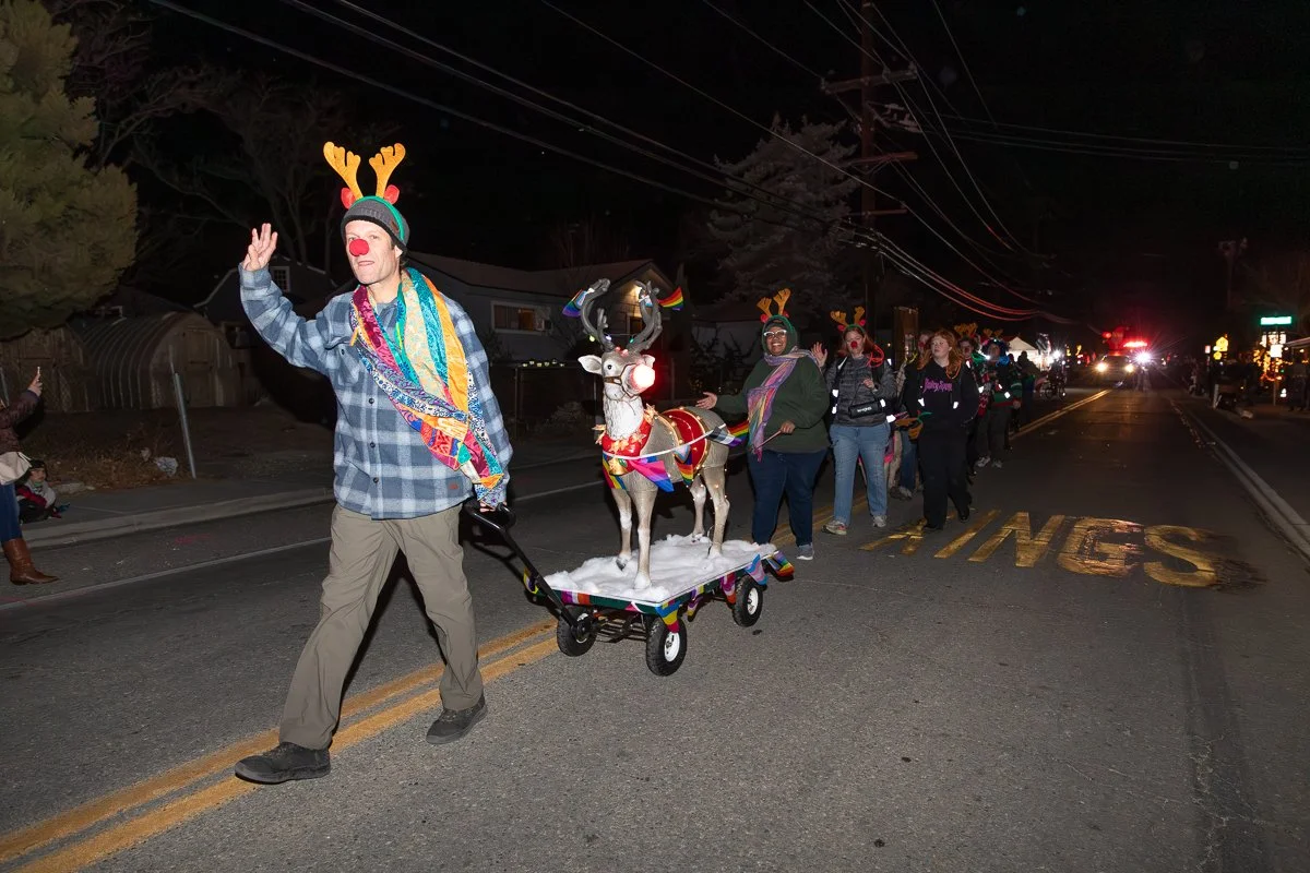 Group of people participating in a night parade dressed in Christmas and reindeer costumes, walking down a street, some with reindeer antlers and red noses, with a small reindeer figure on a decorated wagon.