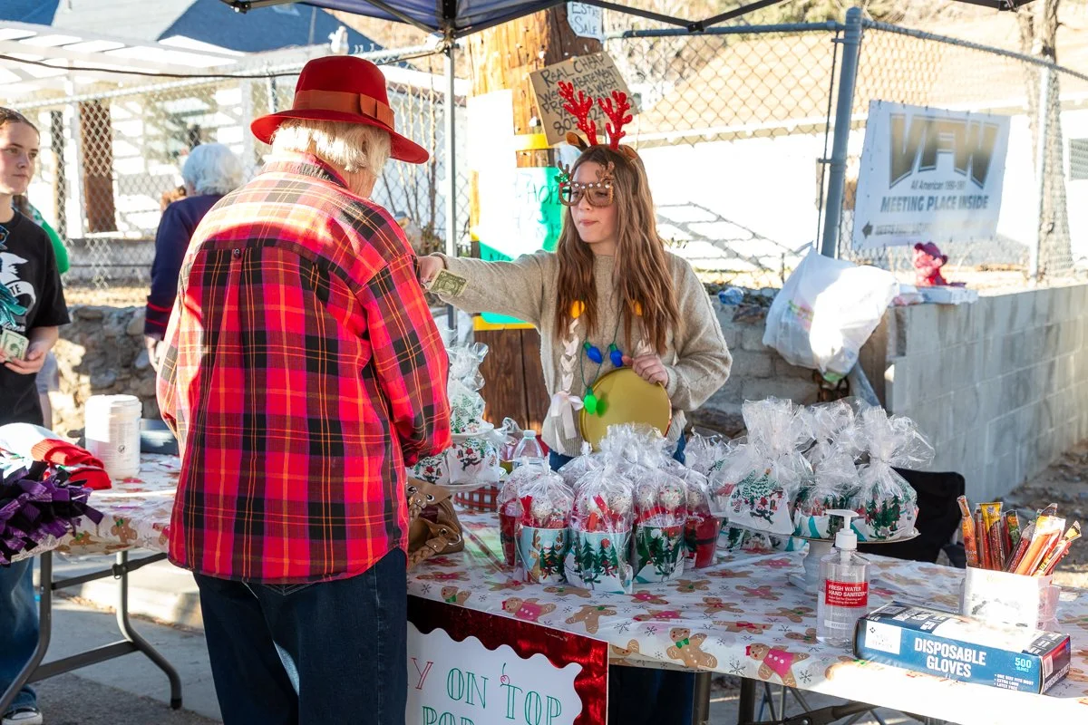A woman dressed as an elf with Christmas lights on her sweater and reindeer antlers accepting money from a woman wearing a red plaid shirt and red hat at a holiday gift stand outside. The stand has wrapped holiday presents and decorations, with a sig