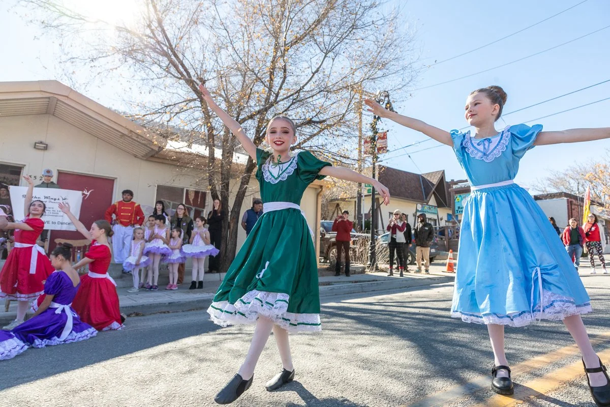 Children performing a dance on a street during a parade, dressed in colorful Victorian-style dresses in green, blue, and red, with spectators watching.