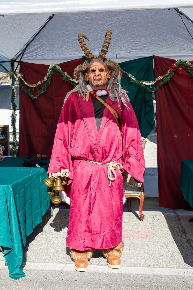 Person dressed in a festive costume with a demonic mask, large horns, and a red robe standing under a holiday decorated tent, holding a bundle of bells.