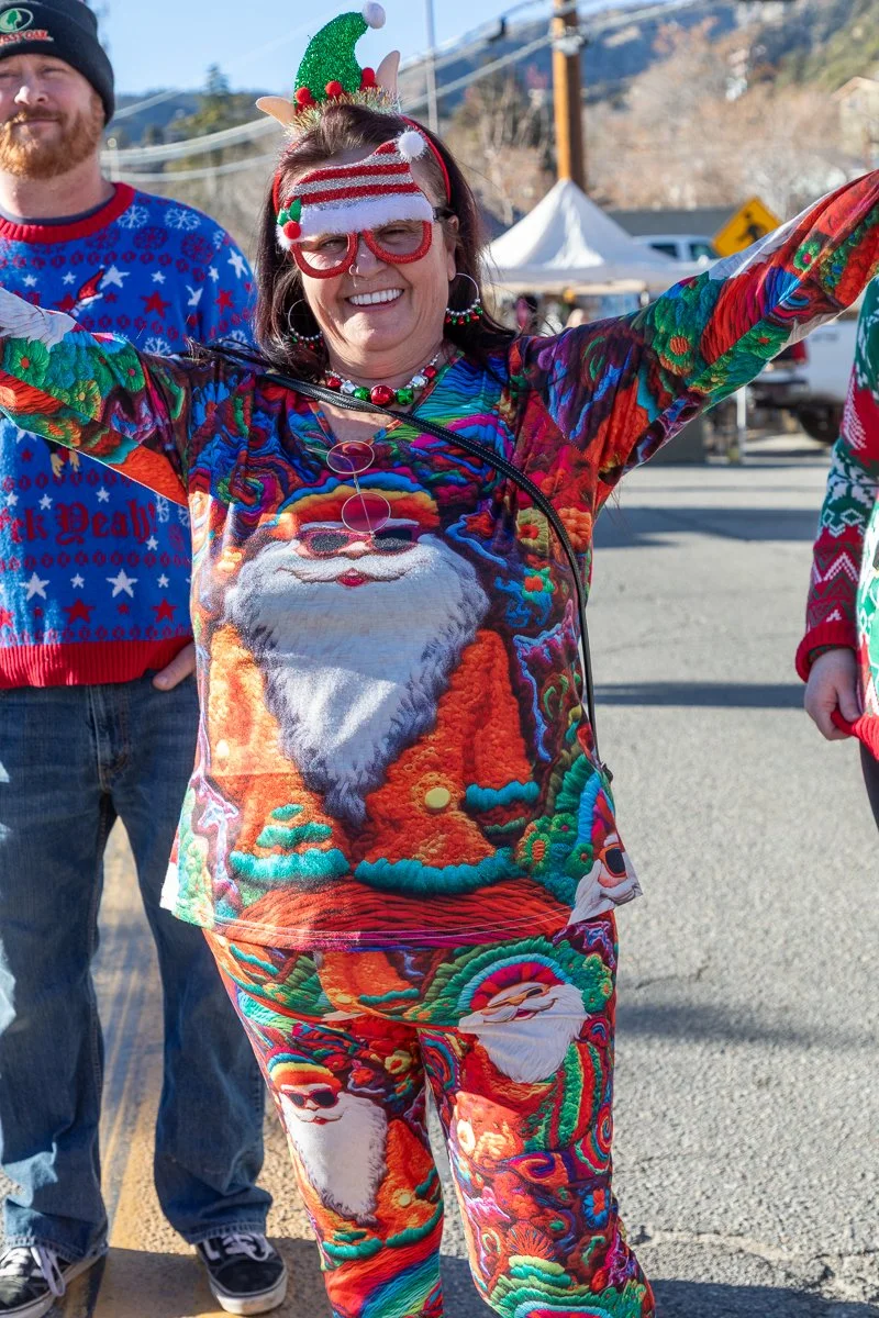 A woman wearing a festive Christmas sweater and pants with Santa Claus designs, a striped Santa hat, and elf ear headband. She is smiling with her arms raised outside on a sunny day, with a man in a holiday sweater and other people and tents visible