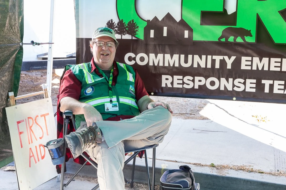 A smiling man sitting in a chair at a community emergency response team event, wearing a green vest and a cap, with a first aid sign and an emergency response banner in the background.