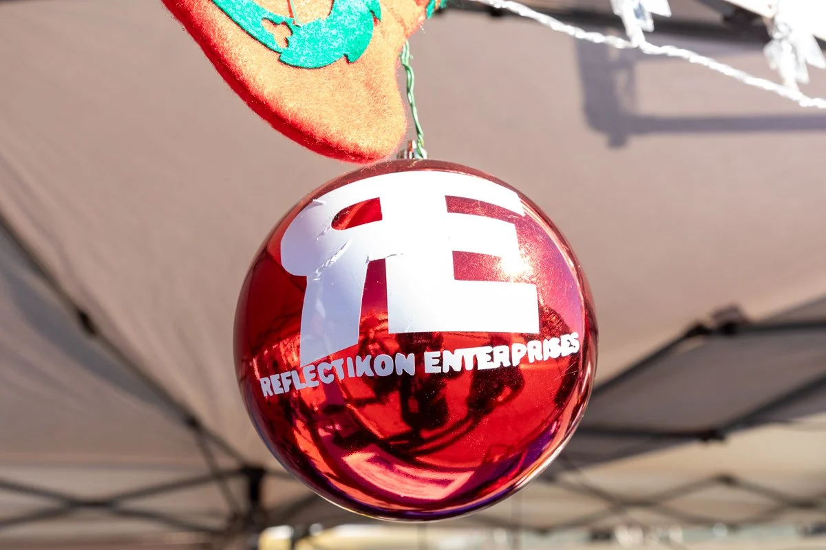 A red, reflective Christmas ornament hangs from a string. The ornament has white lettering that says "REFLECTION ENTERPRISES" and a large white logo with the initials "RE." In the background, there is a beige tent canopy and a few blurred objects.