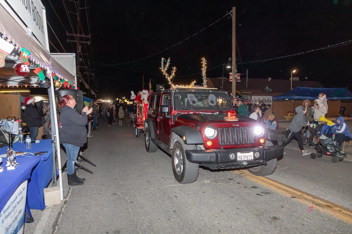A nighttime street scene with a decorated Jeep with reindeer antlers and lights, part of a holiday parade or celebration, with people gathered around on both sides of the street.